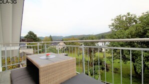 A balcony with a wicker table and a cushion, overlooking a green landscape with trees and houses. - Overath Luxus Apartments (Overath)