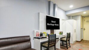 Habitación de hotel con una televisión de pantalla plana mostrando “Hotel Heritage Home”, un escritorio con sillas, un sofá de cuero marrón y una alfombra roja.