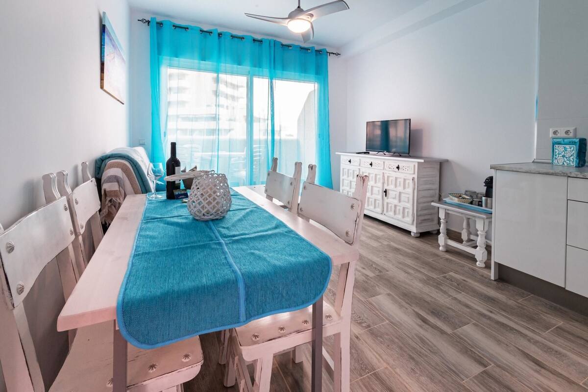 Ein Essbereich mit Tisch, der mit einer blauen Decke bedeckt ist, weißen Stühlen, einem weißen Schrank mit Fernseher und einem kleinen Tisch mit Korb.