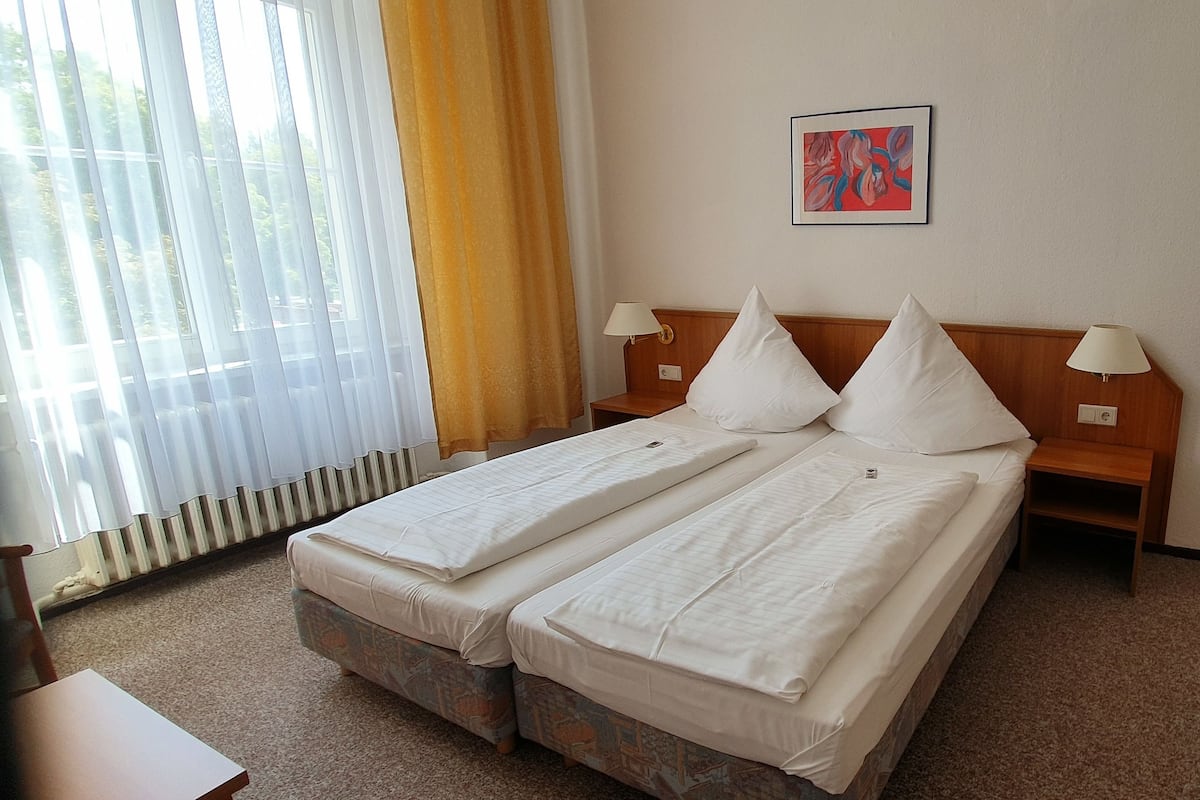 Habitación de hotel con dos camas individuales, cabecero de madera, una mesita, una silla, un radiador y una ventana con cortinas.