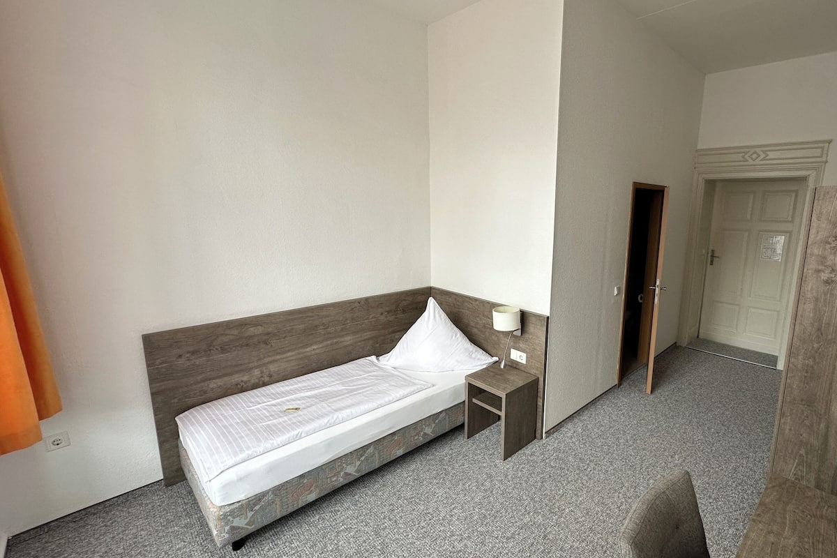 Una cama individual con cabecera de madera, una mesita de noche con lámpara, una puerta que da a otra habitación y suelo cubierto de alfombra.