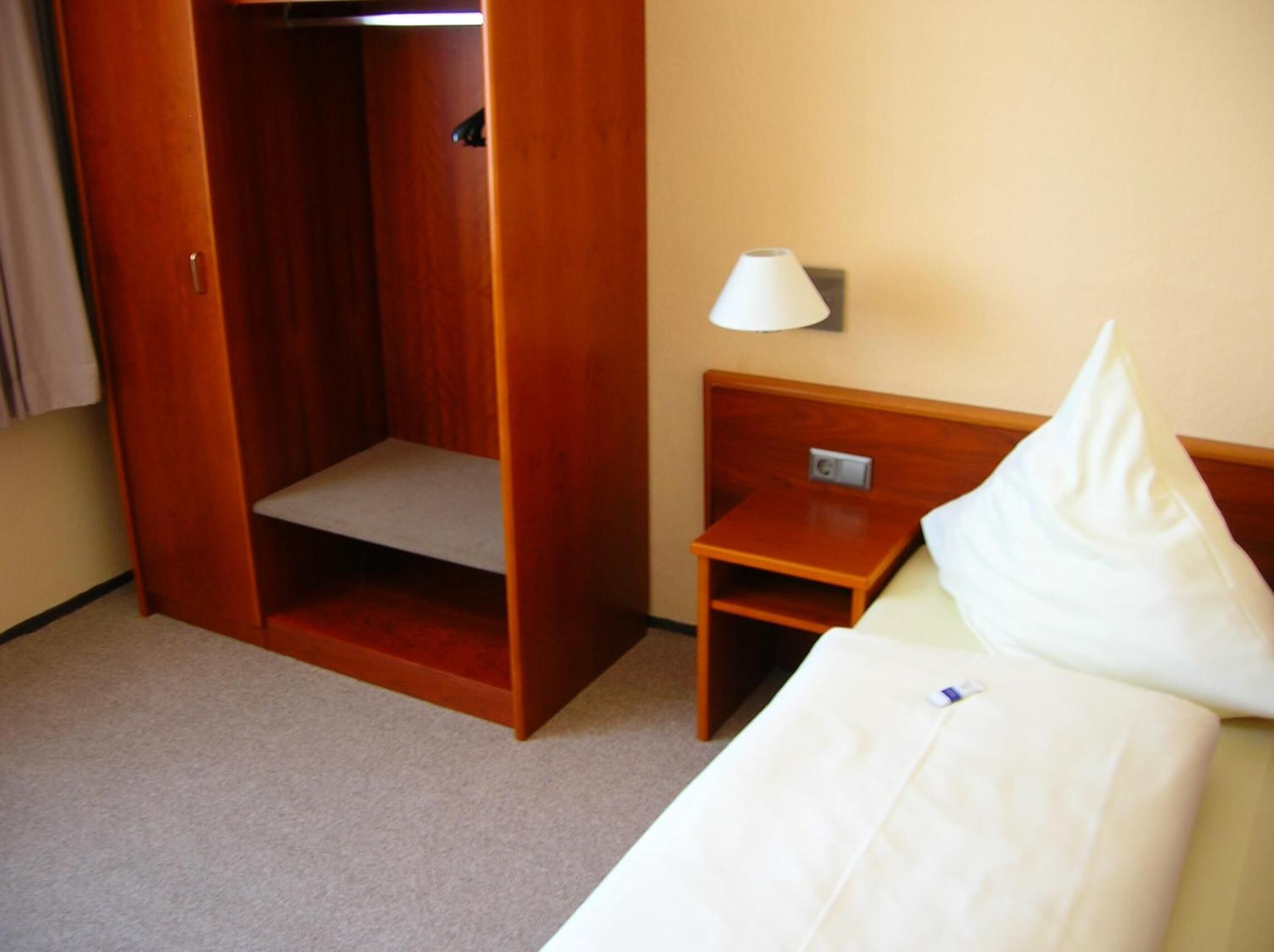 A hotel room with a wooden wardrobe, a bedside table with a lamp, and a bed with a pillow.