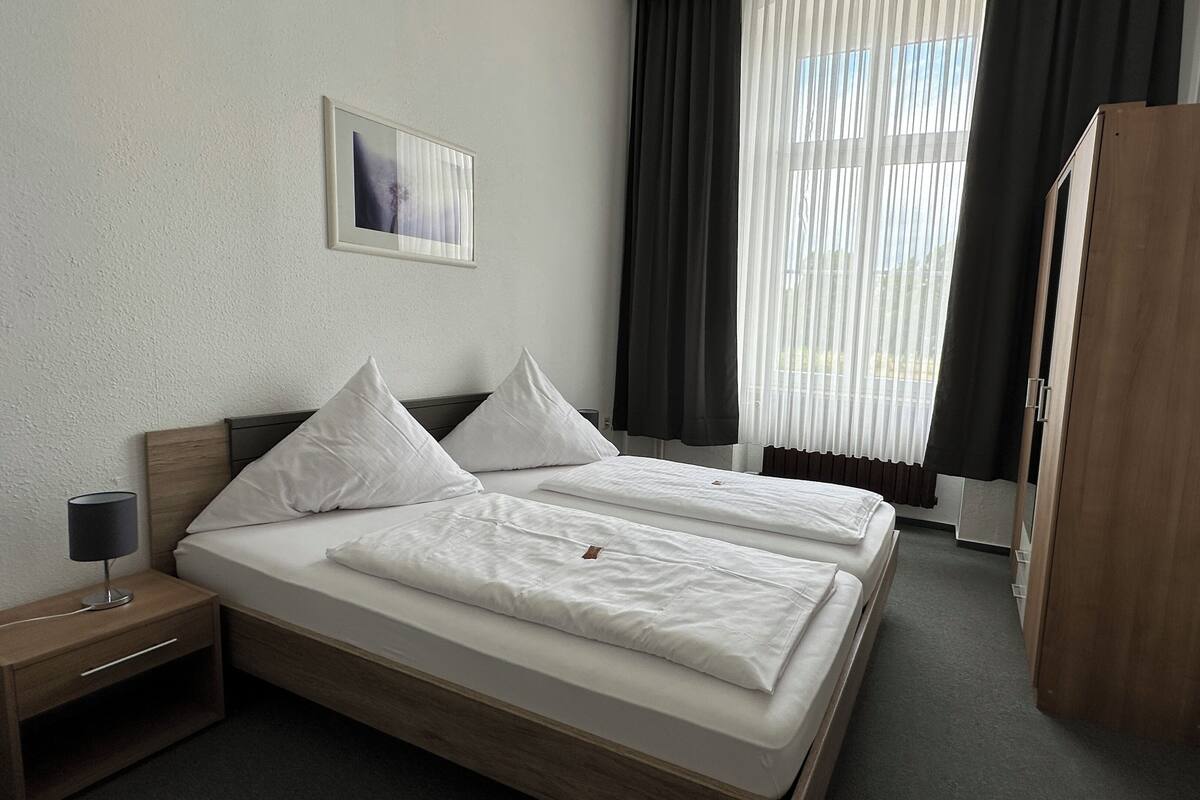 Habitación de hotel con dos camas, una mesita de noche, una lámpara, una ventana con cortinas y un armario de madera.