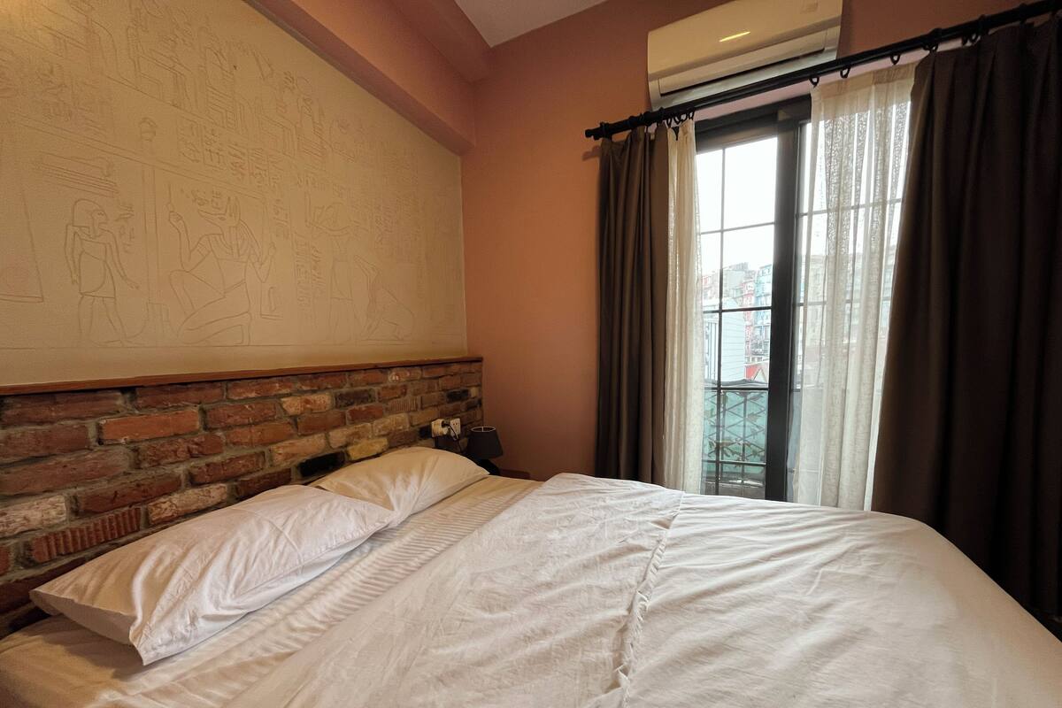 Una habitación de hotel con cabecera de ladrillo, un ventanal con cortinas y una cama con sábanas blancas.