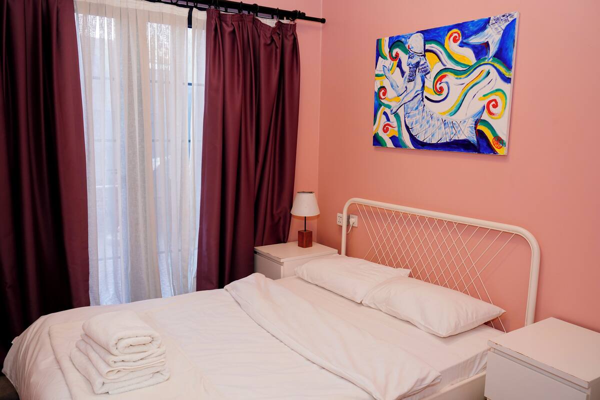 Una cama bien hecha con sábanas blancas, una mesita de noche con lámpara y un cuadro abstracto de colores en la pared.