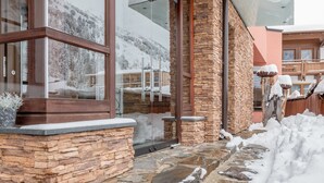 Fachada de piedra con puerta de cristal que da a un interior de madera, nieve en el suelo y vistas a la montaña.