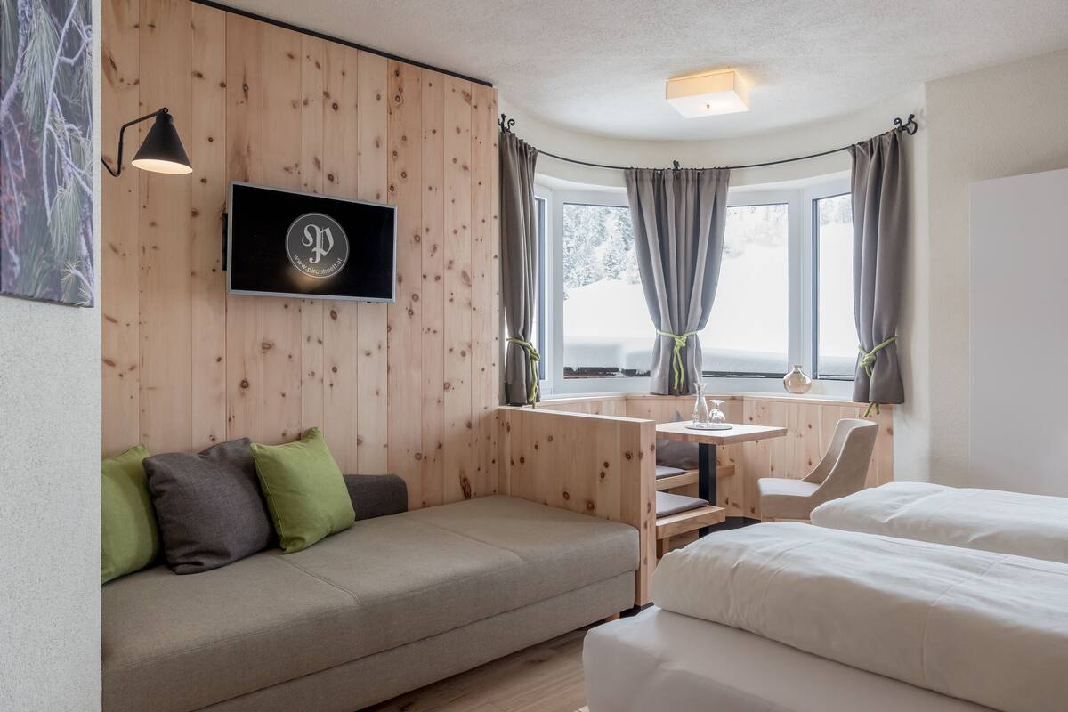A cozy room with a wooden wall, a flat-screen TV, a sofa with cushions, a desk with a chair, and two beds.