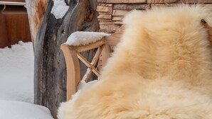 Una silla de madera rústica con un cubre de piel, una pared de piedra y esculturas cubiertas de nieve en un entorno invernal al aire libre.