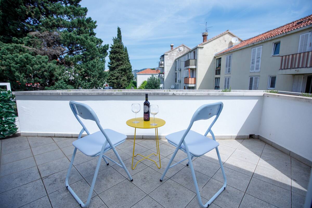 A balcony with two chairs and a small round table, a bottle of wine, and a view of buildings and trees.