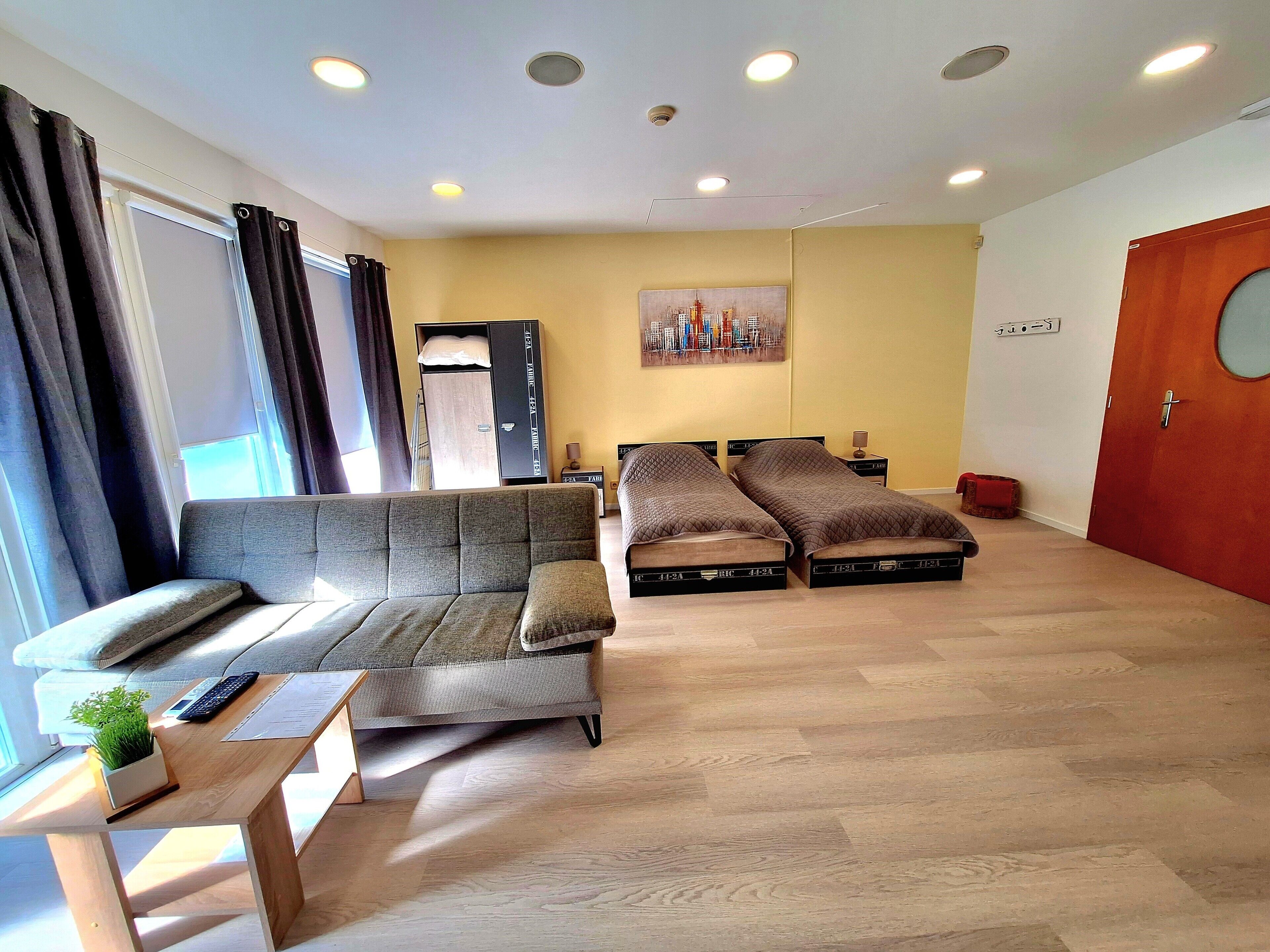 Una sala moderna con un sofá, dos camas, piso de madera, un cuadro en la pared y una puerta con un ventanal redondo.