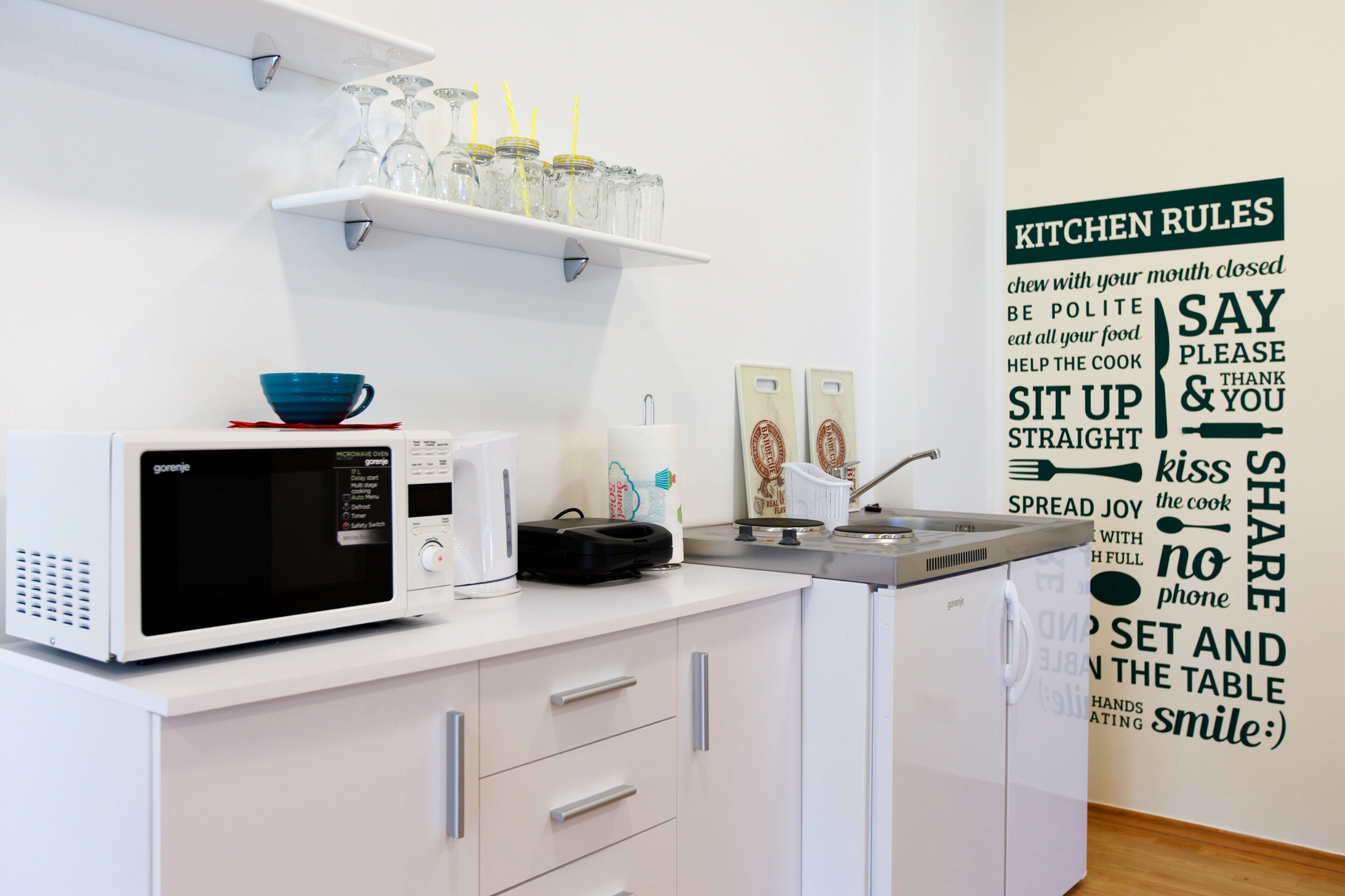 A modern kitchen with a microwave, kettle, and kettle on the counter. There is a shelf above the counter holding glasses and a bowl.