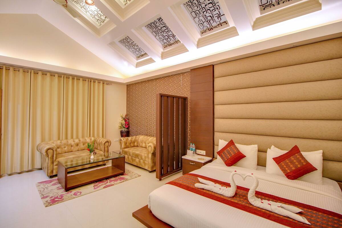 A hotel room with a bed, two sofas, a glass coffee table, a nightstand, and a decorative wall panel.