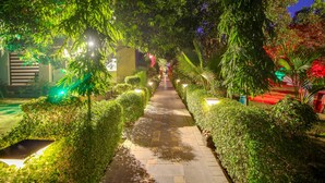 A nighttime garden pathway with neatly trimmed hedges and stone paving, illuminated by soft lighting. - Maulik Mansion Resort (Ramnagar)