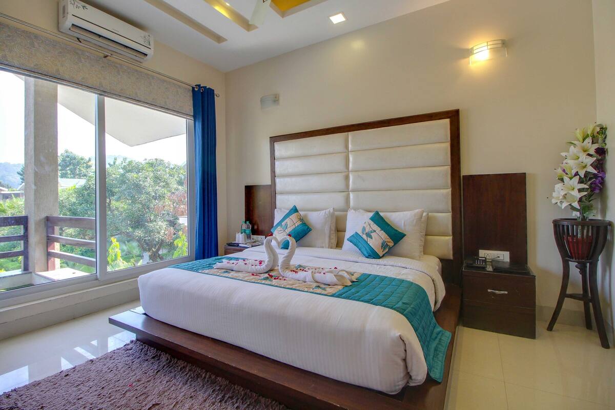 A hotel room with a large bed, a nightstand, a small table, and a window with a view of greenery.