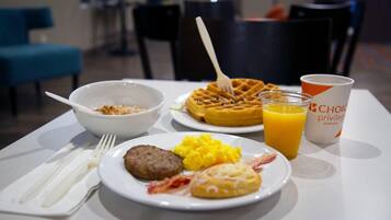 A breakfast table with a waffle, scrambled eggs, sausage, and a glass of orange juice.