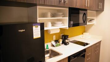 A compact kitchen with a black refrigerator, a microwave, a sink, and a dishwasher. There are shelves above the sink holding plates and cups.