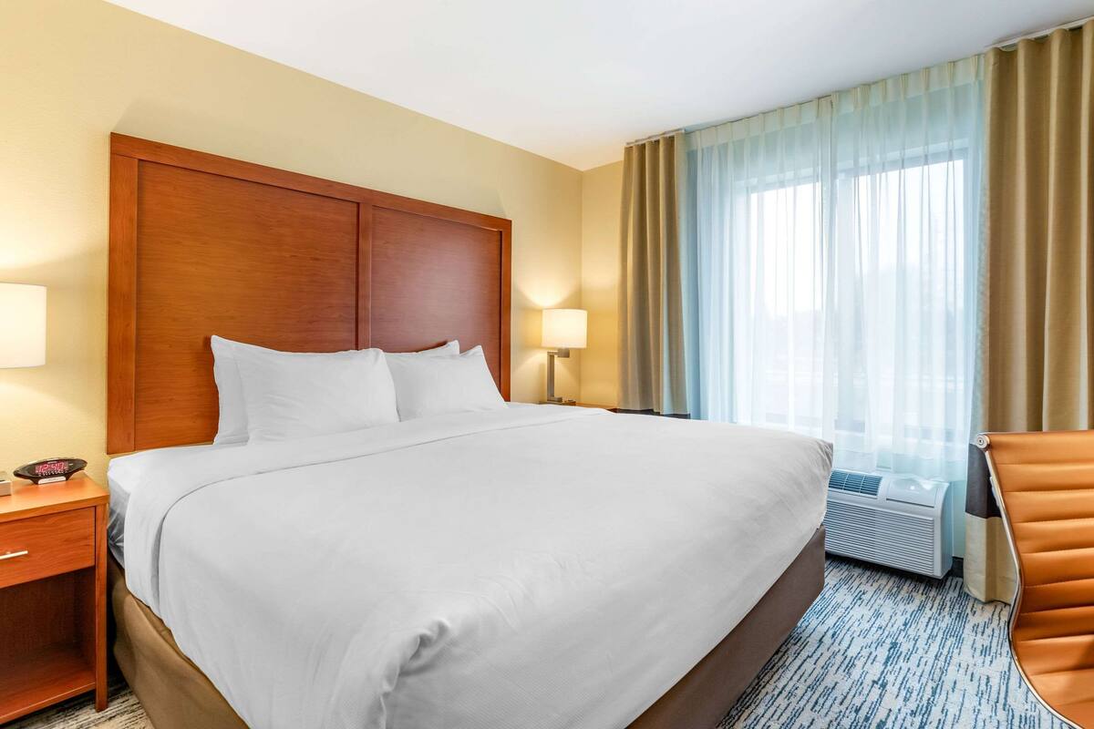 A hotel room with a large bed, wooden headboard, two bedside tables with lamps, a window with curtains, and a brown leather chair.