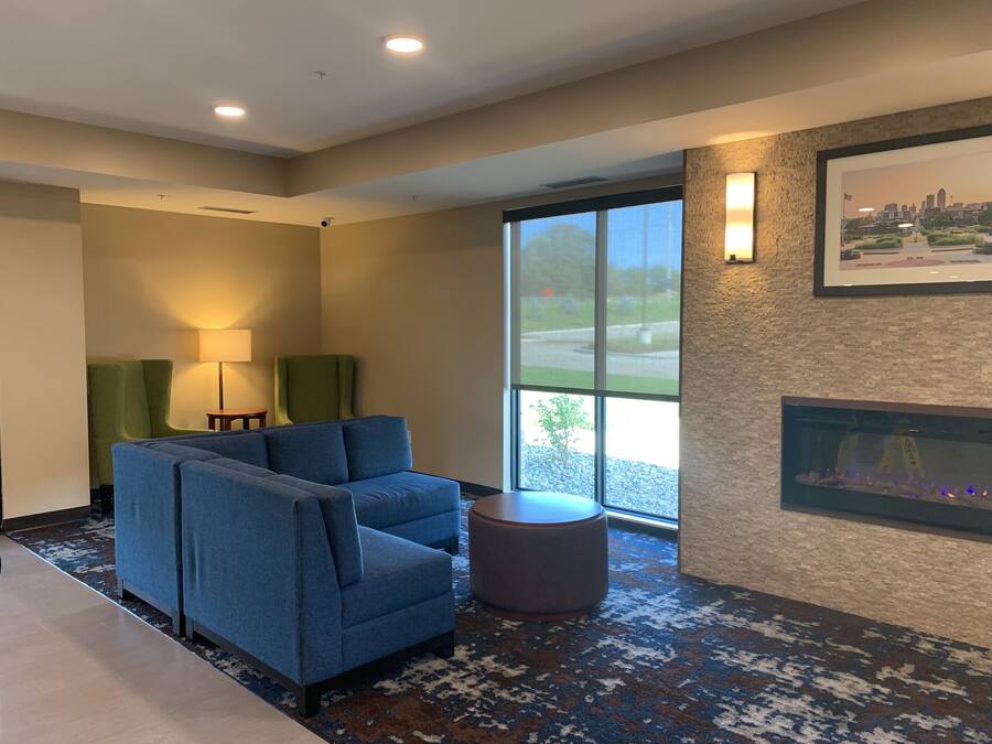 A modern hotel room with a blue sofa set, a small round ottoman, a fireplace, and a large window offering a view of the city skyline.