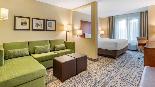 A hotel room with a green sofa, a bed, a small table, and a mirror. There are three framed pictures on the wall and a window with curtains. - Comfort Inn & Suites West Des Moines (West Des Moines)