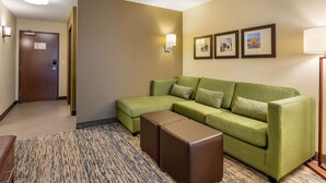 A hotel room with a green sectional sofa, a brown ottoman, a patterned rug, and framed pictures on the wall. - Comfort Inn & Suites West Des Moines (West Des Moines)