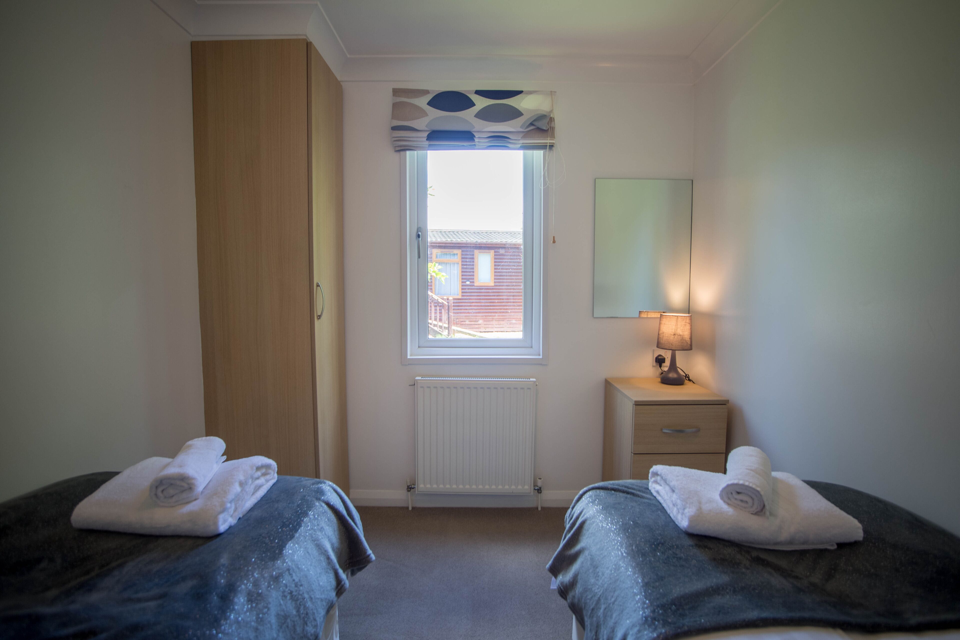 A room with two beds, a window with blinds, a radiator, a bedside table with a lamp, and a mirror on the wall.