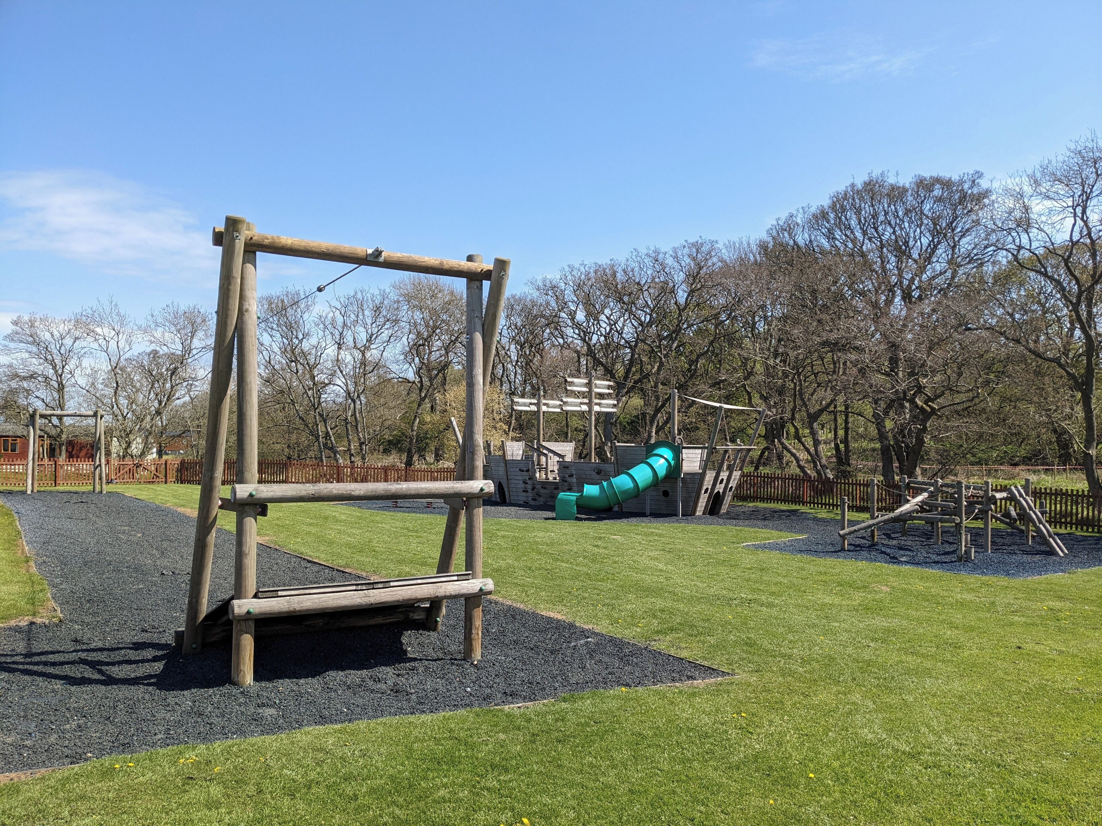 A playground with a wooden swing set, a slide, and a fenced area with trees in the background.