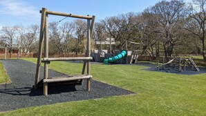 A playground with a wooden swing set, a slide, and a fenced area with trees in the background. - Felmoor Park Northumberland Lodge With Hot Tub - 4 (Morpeth)