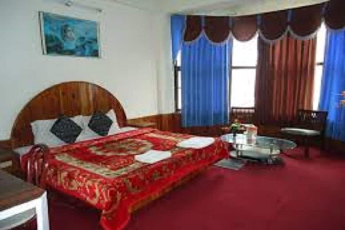 Habitación con una cama grande, una mesita pequeña y una silla. Se ve una ventana con cortinas.