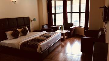 A hotel room with a large bed, a desk, and a chair. There are windows with curtains, a wall-mounted TV, and a ceiling with wooden beams.