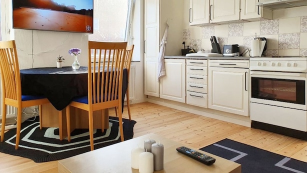 A compact kitchen with white cabinets, a stove, and a sink. There's a dining area with a table and chairs, and a TV mounted on the wall. - Struer Town Apartment (Struer)