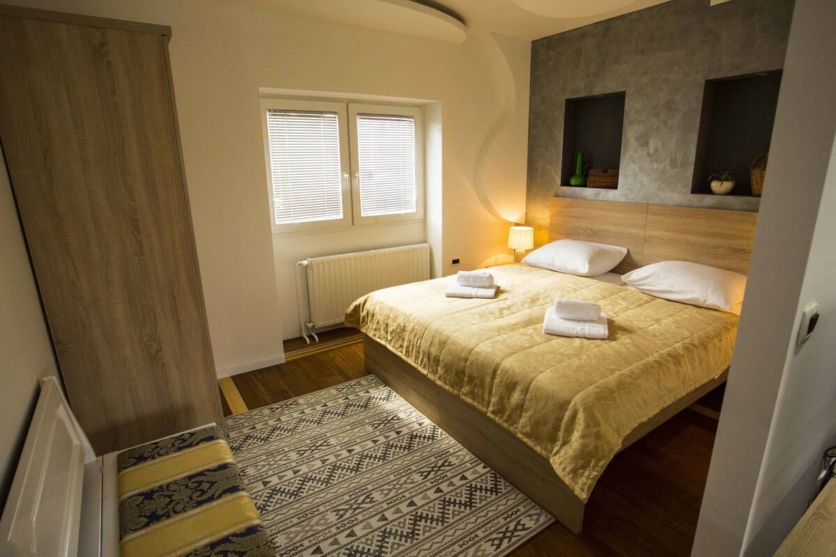 A bedroom with a bed, a window with blinds, a bedside lamp, a wooden headboard, and a patterned rug.