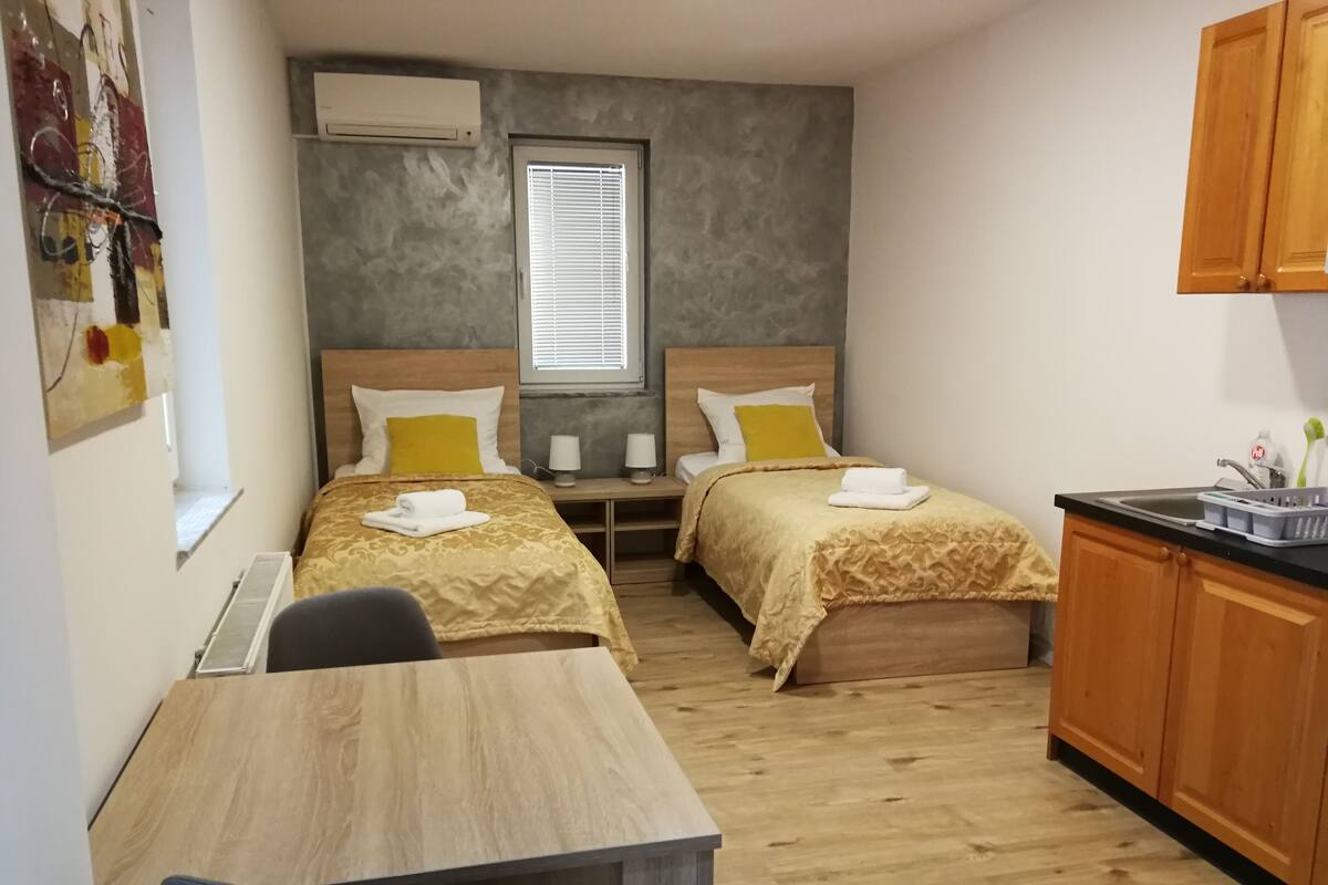 A compact hotel room with two beds, a small kitchenette, a wooden desk, and a window with blinds.