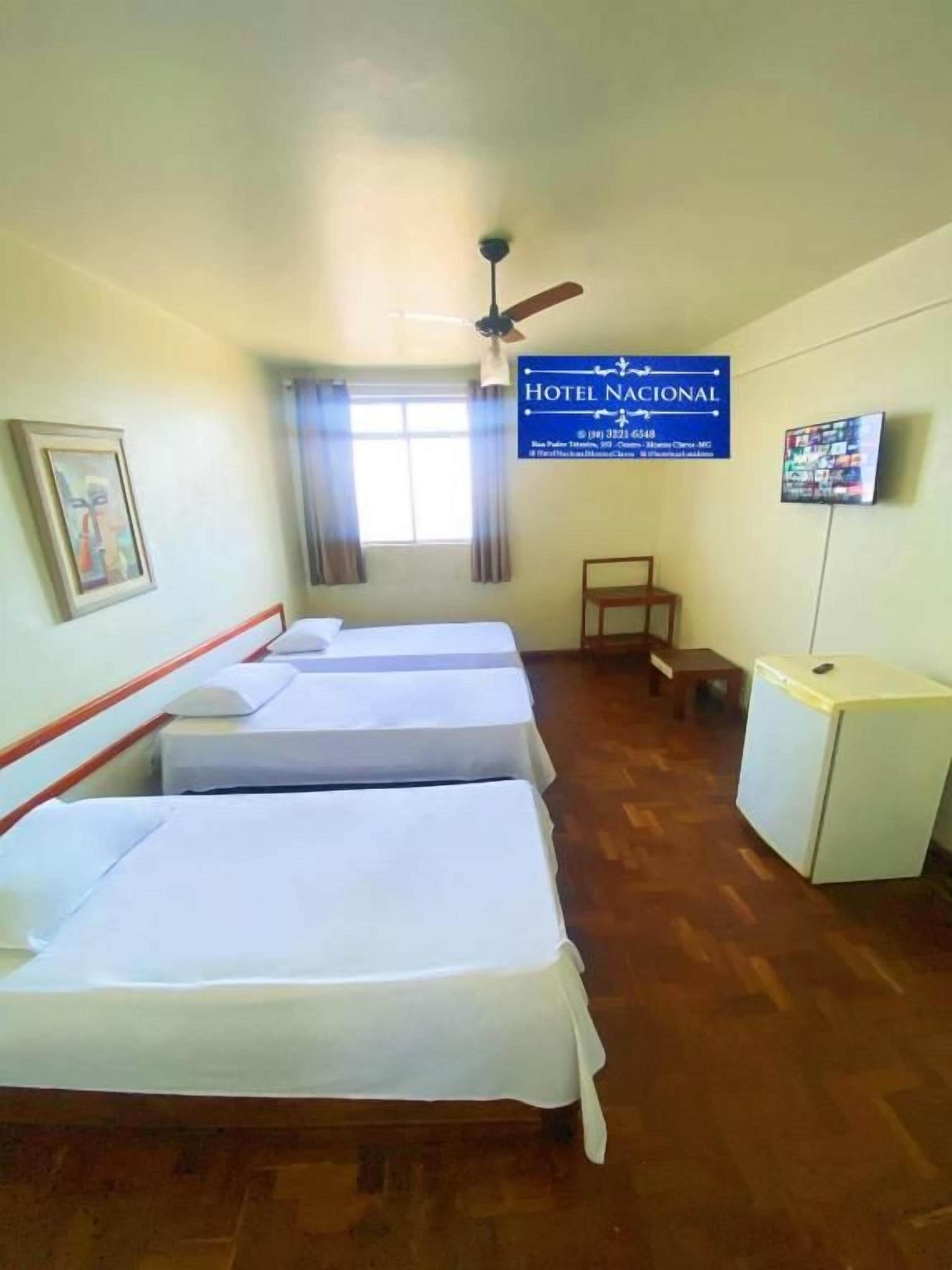 Quarto de hotel com três camas, uma escrivaninha, televisão e ventoinha no teto.