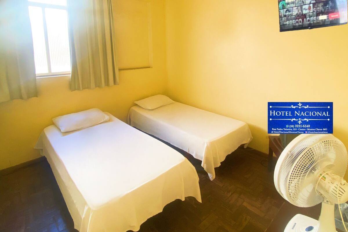 Hotellihuone, jossa on kaksi erillistä sänkyä, kattotuuletin, ikkuna verhoilla, seinälle kiinnitetty kyltti sekä pöytä, jossa on tuuletin ja jokin laite.