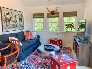 A living room with a sofa, a wooden chair, a patterned rug, a metallic coffee table, and a red dice cube.
