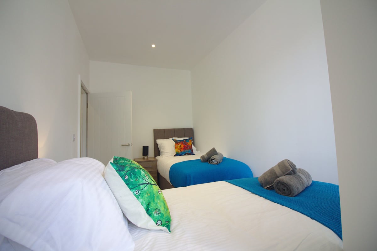 Une chambre d’hôtel avec deux lits, l’un avec du linge de lit blanc et l’autre avec du linge de lit bleu. Il y a des serviettes sur les lits et une petite table avec une lampe.