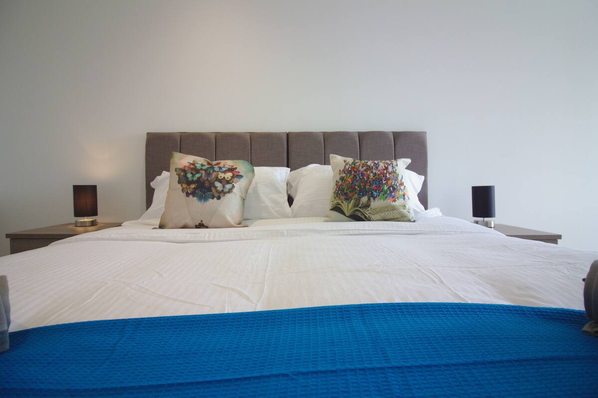 Un lit bien fait, recouvert d’une couverture bleue, avec deux coussins décoratifs et deux lampes de chevet posées sur une table de nuit.