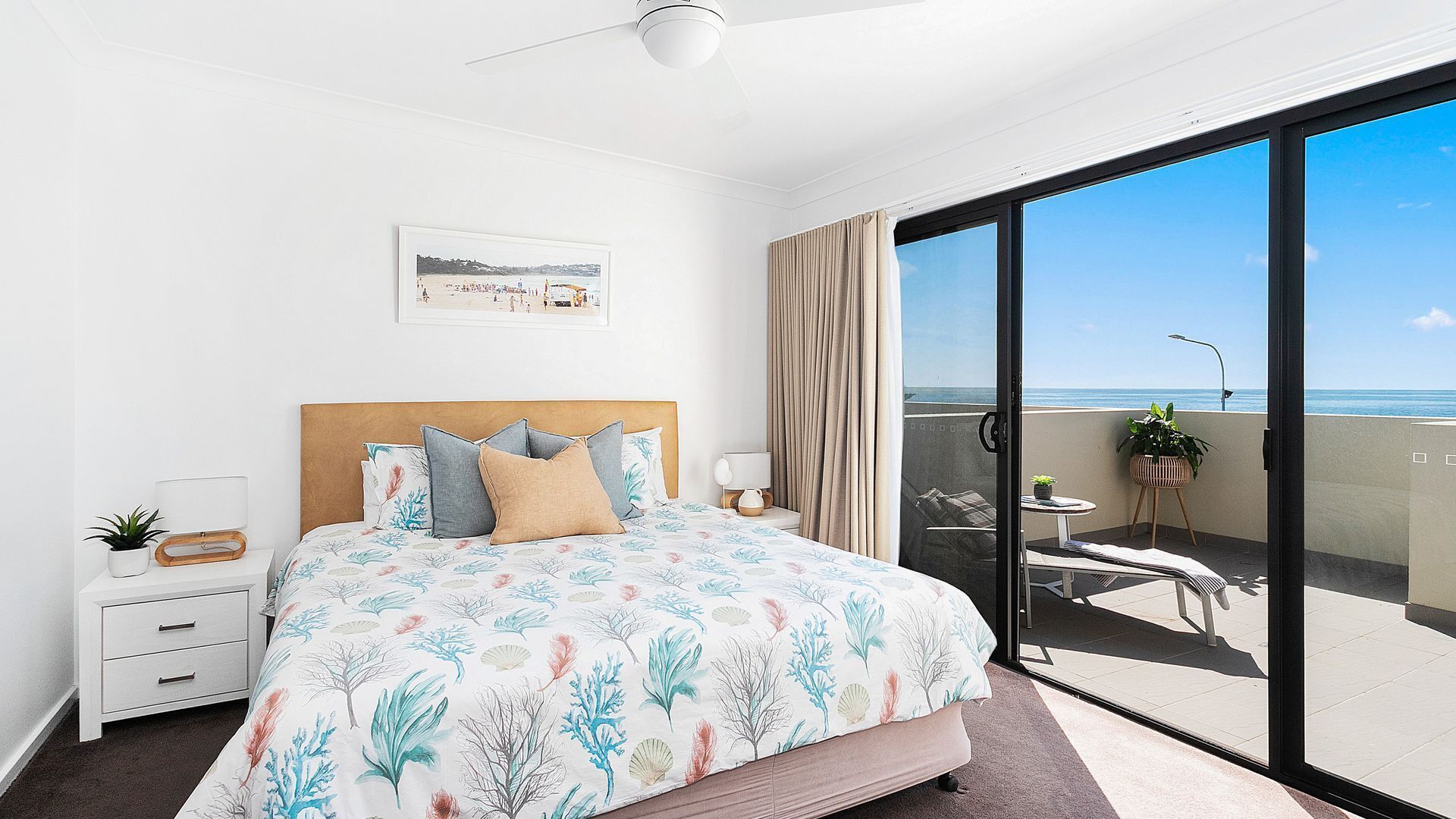 A bedroom with a bed, a bedside table, a lamp, a framed picture, and a balcony with a view of the sea.