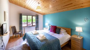 A bedroom with a wooden ceiling, a large bed with a blue bedspread, a bedside table with a lamp, and a window with curtains. - Woodpecker Lodge With Hot Tub Near Cupar, Fife (Cupar)