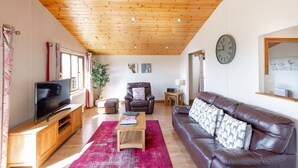 A living room with a wooden ceiling, a television on a wooden cabinet, a brown leather sofa, a red rug, and a clock on the wall. - Woodpecker Lodge With Hot Tub Near Cupar, Fife (Cupar)