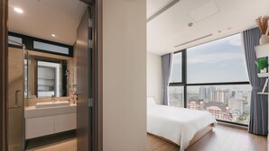 Una habitación de hotel moderna con un ventanal amplio que ofrece vistas a la ciudad, un baño independiente con tocador y una cama con edredón blanco.
