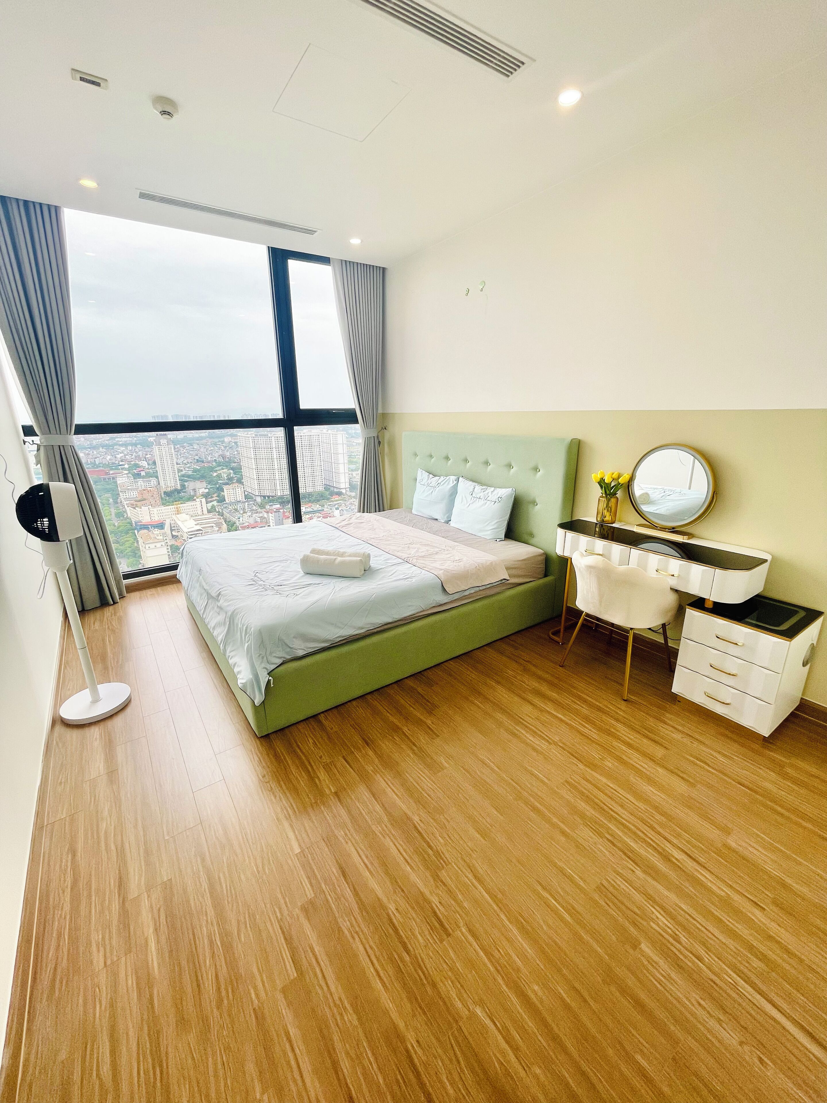 A modern bedroom with a large bed, a bedside table with a mirror, and a view of the city through large windows.