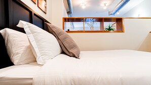 Un letto rifatto con cura, con biancheria bianca e beige, una testiera in legno e una finestra che si affaccia sul cielo e sulla vegetazione.