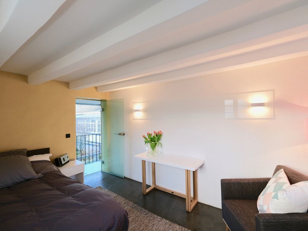 A modern hotel room with a bed, a sofa, a small table with a vase of flowers, and a balcony door.