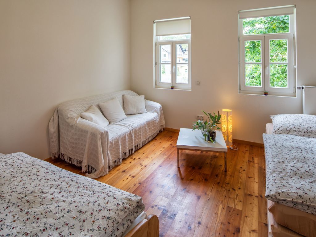 A cozy room with a wooden floor, a white sofa, a small table with a plant, and two windows providing natural light.