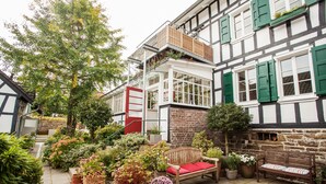 A traditional half-timbered building with a red door, a wooden bench with red cushions, and various potted plants. - Haus in Bewegung (Neunkirchen-Seelscheid)