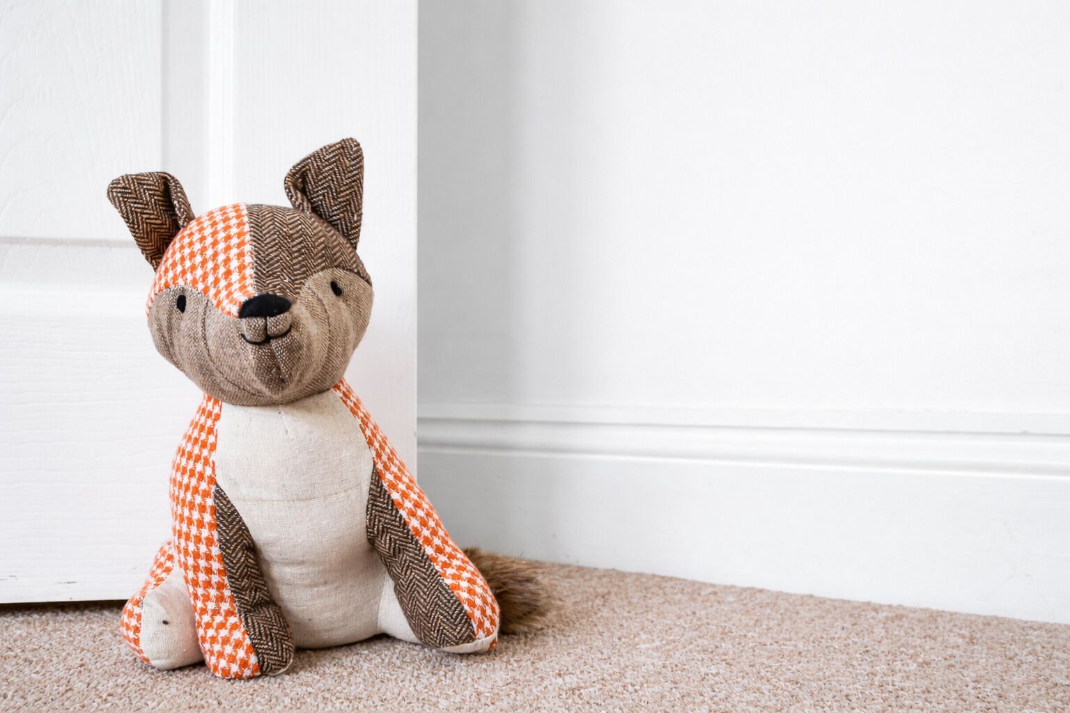 A plush toy with a checkered pattern sitting on a carpeted floor against a white wall.