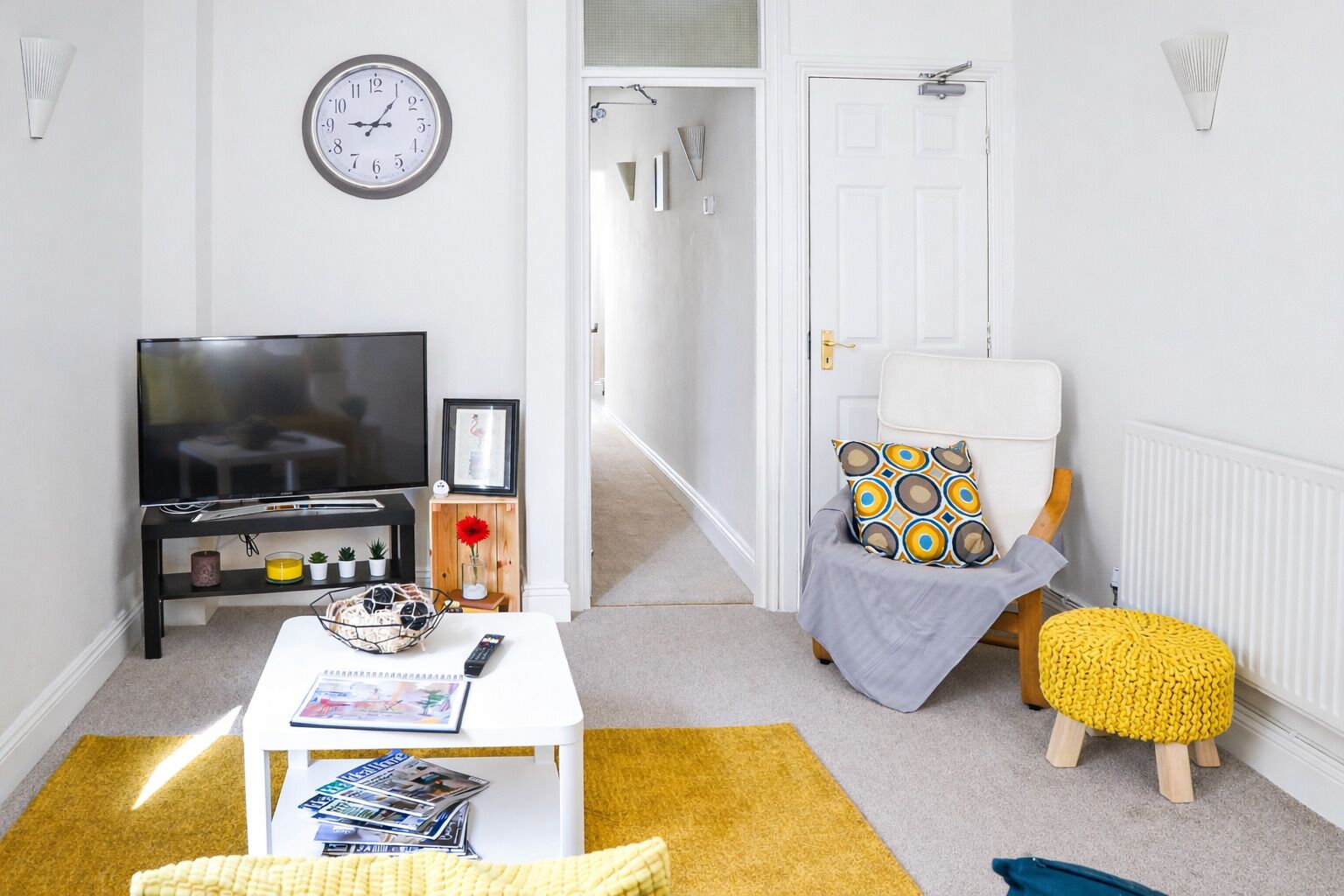 A modern living room with a flat-screen TV, a coffee table with magazines, a yellow rug, a white armchair with patterned cushions, and a small wooden stool.
