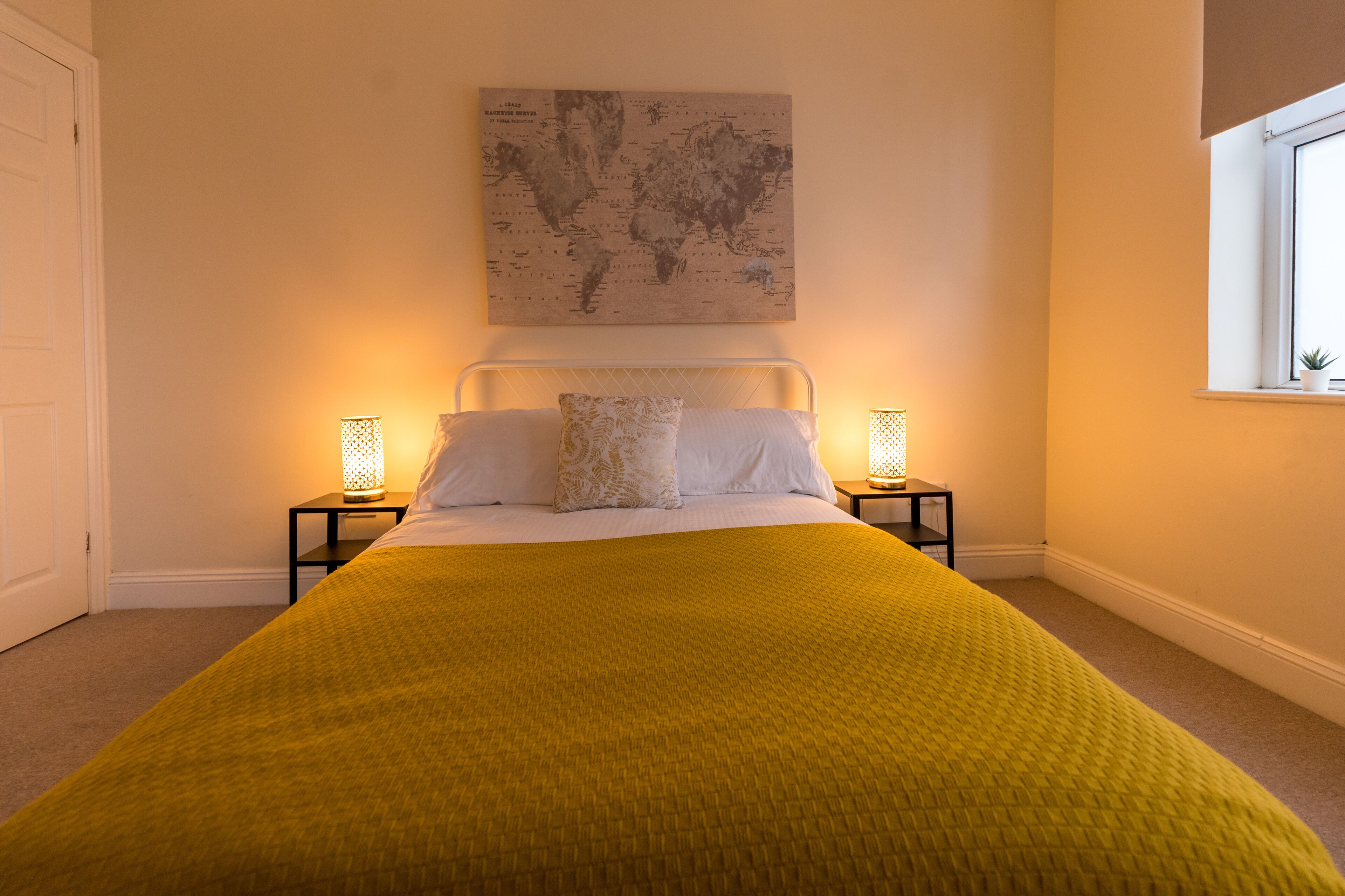 A neatly made bed with a yellow blanket, two bedside tables with lamps, a world map artwork on the wall, and a window with a plant.