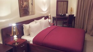 Une chambre d’hôtel avec un lit en bois, des oreillers blancs, une couverture rouge, un bureau avec une lampe, un miroir et un tableau au mur.